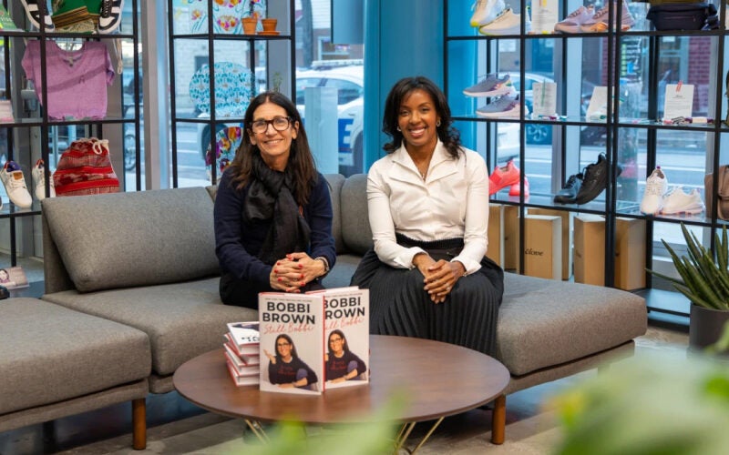 Two people sitting on a sofa in a store setting, with copies of a book titled "Bobbi Brown" on a table in front of them. The background features shelves with shoes and other items.