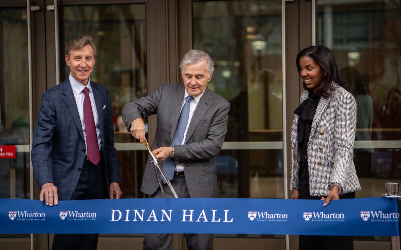 A few individuals are participating in a ribbon-cutting ceremony at Dinan Hall. One person is cutting a ribbon, and they are all dressed in formal attire.