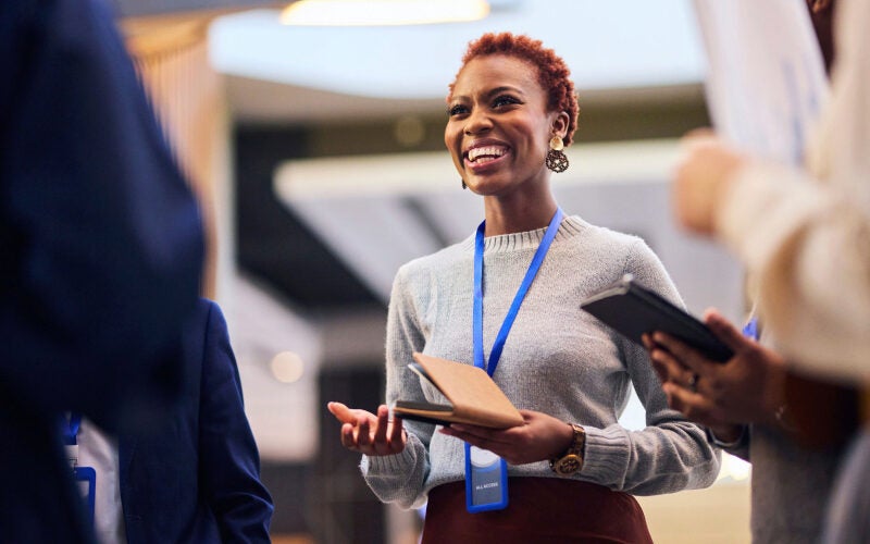 A person smiling and talking in a professional setting, holding a notebook, with others around them. They are wearing a name badge on a blue lanyard.