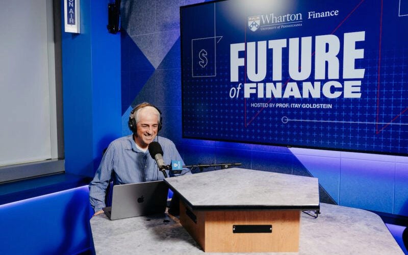 A person sitting at a podcast studio desk with a microphone, laptop, and headphones. Background displays "Future of Finance" text.