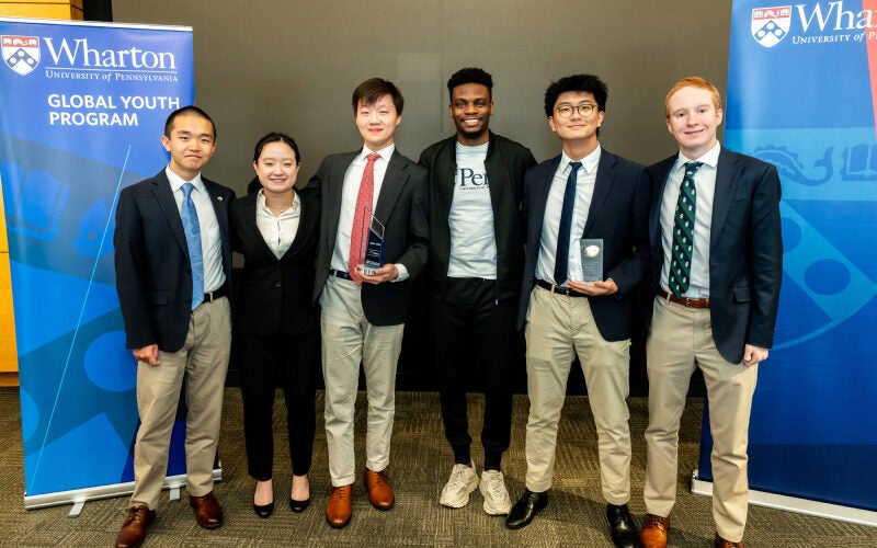 A small group of people in formal attire posing for a photo, standing in front of banners for the Wharton Global Youth Program at the University of Pennsylvania.