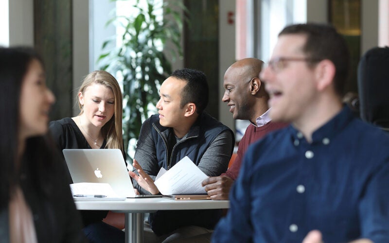A small group of people sitting around a table engaged in a discussion. One person is using a laptop, while others are holding documents.