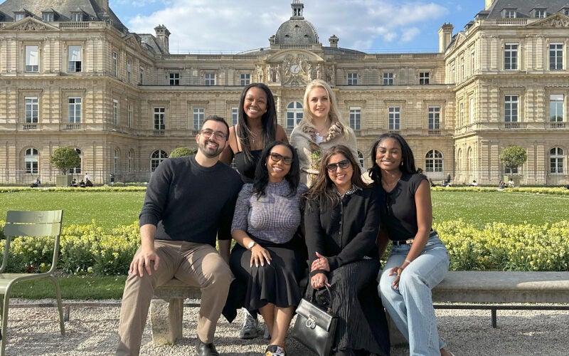 A group of people smiling and posing in front of a large, historic building with manicured gardens.