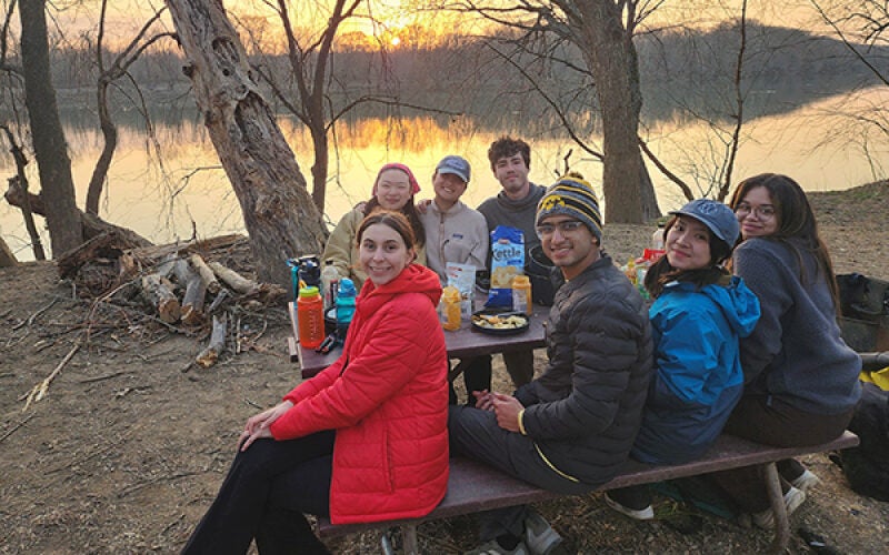 A group of people sitting at a picnic table near a body of water, with a sunset in the background. They are enjoying snacks and drinks outdoors.