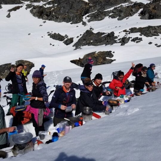 A group of smiling people relax for a break with food and drink in a snowy mountainous area.