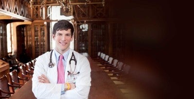 A doctor wearing a white coat and stethoscope stands confidently with arms crossed in a large, ornate conference room.