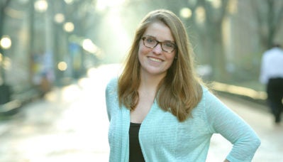 A person with long hair and glasses smiling, standing outdoors on a sunlit path lined with trees.