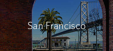 A view of the San Francisco Bay Bridge framed by a brick arch, with palm trees and waterfront buildings visible in the foreground. The text "San Francisco" is overlayed.