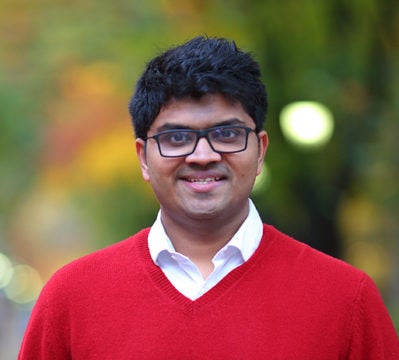 Headshot of a person wearing glasses and a red sweater, smiling, with a blurred background of greenery and lights.