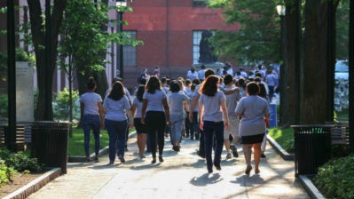A large group of people wearing matching shirts walking along a tree-lined path on a sunny day, possibly a campus or park setting.