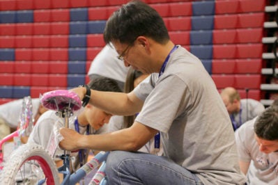 A person assembling a children''s bicycle in an indoor setting with colorful padded walls in the background. Several others are engaged in similar activities.