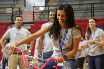 A person wearing a Wharton School t-shirt is joyfully riding a decorated pink bicycle indoors, with a small group of people in similar shirts watching and smiling.