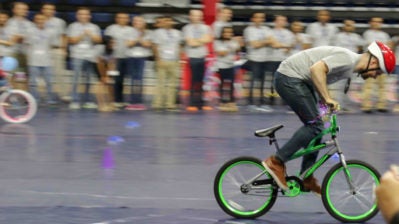 A person is riding a bicycle indoors while wearing a red helmet. A group of people in the background appears to be observing the activity.