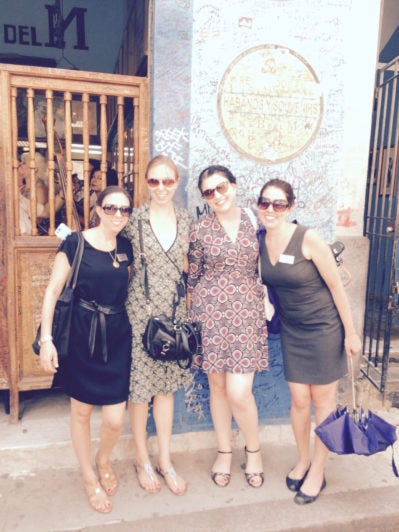 A group of four people standing in front of a blue wall covered in graffiti, smiling and posing for a photo. They are wearing summer dresses and sunglasses. The setting appears to be an entrance to a shop or café.