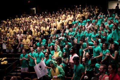 A large group of people in a theater, divided into two sections. One group is wearing yellow shirts and the other is in green shirts, showing enthusiasm and holding signs.