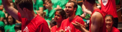 A group of people wearing matching red shirts are cheering and smiling, with others in green shirts in the background, suggesting a lively team event or gathering.