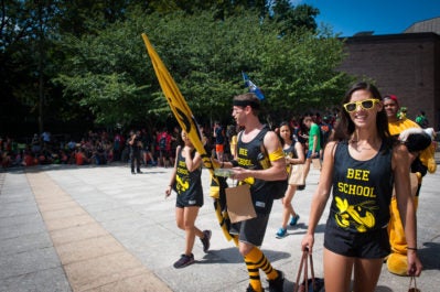 A group of people in matching "Bee School" outfits and accessories are participating in a festive outdoor event. They are holding a yellow flag and walking near a crowd under a sunny sky.