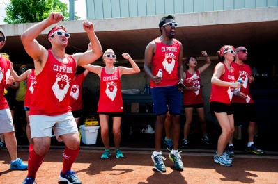A group of people wearing matching red tank tops with "CI PRIDE" and a lion emblem, engaged in an energetic outdoor activity. They are wearing sunglasses and appear to be cheering or exercising together in a sunny setting.