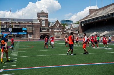 A group of people playing a sport on a green field surrounded by a stadium with stands. The weather is sunny with a clear sky.