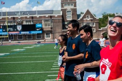 A group of people laughing and enjoying a sunny day on a sports field, with a brick building and scoreboard in the background.