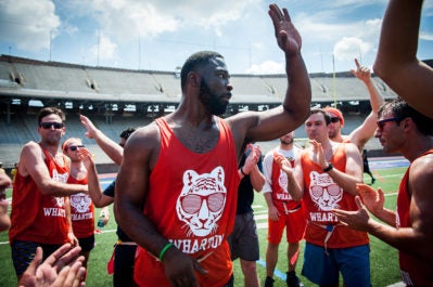 A group of people on a sports field, wearing matching orange tank tops with a tiger design and "Wharton" text, appear to be participating in a team event.