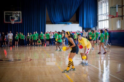 A group of people playing dodgeball in a gymnasium. Some wear green shirts, while others wear a mix of costumes. Brightly colored balls are in play.